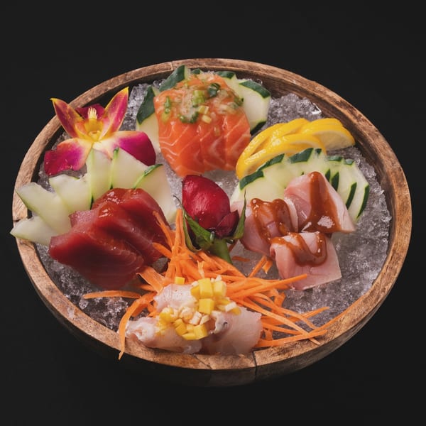 A wooden bowl of Neveyah Sushi and Thai sashimi on ice, garnished with cucumber, lemon, and an orchid.
