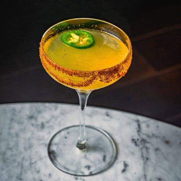 A yellow cocktail with a chili-salted rim and a jalapeño slice, on a marble table.